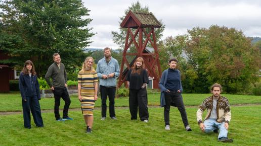 sju personer står på en gräsmatta framför en klockstapel och me utsikt över värmländska Fryken-landskapet.
