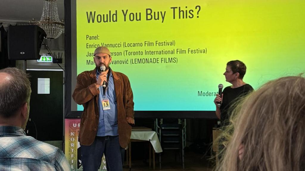 Man med mikrofon framför publik och skärm mad texten Would you buy this?